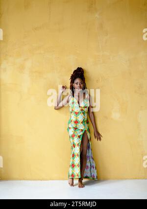 Schöne afrikanische junge Erwachsene Frau, die ein langes Kleid mit hohem Schlitz trägt, in die Kamera blickt, barfuß stehend, auf eine gelbe Wand gelehnt mit c Stockfoto