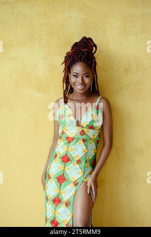 Die attraktive junge afrikanische schwarze Frau, die ein mehrfarbiges Kleid mit hohem Schlitz trägt, in die Kamera blickt und lächelt, isoliert auf einer gelben Wand b Stockfoto