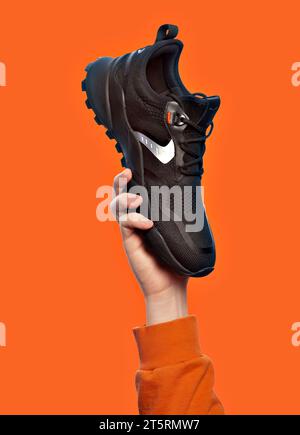 Moderner schwarzer Sneaker, gehalten von der Hand eines Mannes, isoliert auf einem leuchtend orangefarbenen Hintergrund. Kreatives minimalistisches Layout mit Sportschuhen. Outlet Marketin Stockfoto