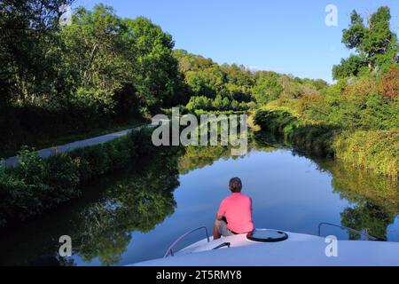 Canal du Nivernais: Bootstour entlang des Nivernais-Kanals mit einem älteren Mann am Bug und Spiegelreflexen von Bäumen in Burgund, Frankreich Stockfoto