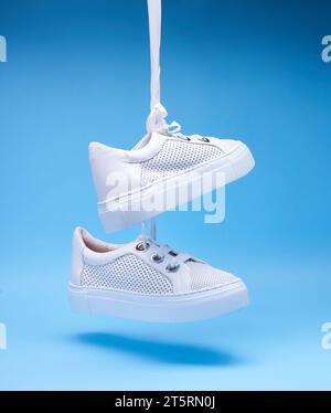 Moderne weiße perforierte Schuhe, die an Schnürsenkeln auf einem blauen Hintergrund hängen. Nahaufnahme. Freizeitschuhe, Sportschuhe. Schuhwerbekonzept Stockfoto
