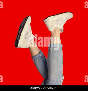 Weibliche Beine tragen trendige weiße Sneakers und Jeans auf einem hellroten Hintergrund. Kreatives Modell für die Werbung für einen Schuhladen. Design für ein f Stockfoto