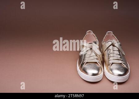 Trendige Sneakers in goldenem Metallic-Design mit zerknitterten Lederdetails und weißen Gummisohlen auf einem abgestuften braunen Hintergrund mit Kopierraum. Modischer Stift Stockfoto