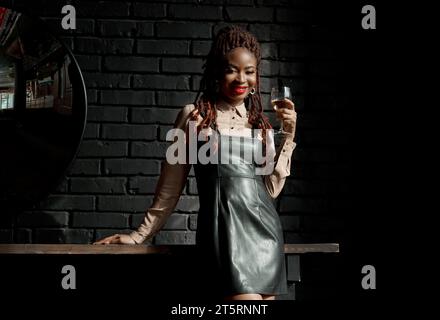 Kühne afrikanische Frau, die luxuriöse Lederschürze trägt, mit einem Glas Wein in der Hand steht und im Restaurant lächelt. Kopierbereich. Cheers, c Stockfoto