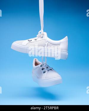 Trendige weiße Sneakers, die an Schnürsenkeln vor einem blauen Hintergrund hängen. Perforiertes Leder, dicke Sohle, breite Schnürsenkel, Metallösen. Fash Stockfoto