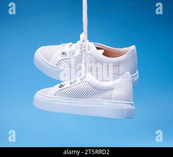 Trendige, legere weiße Sneakers, die vor einem blauen Hintergrund hängen. Perforiertes Leder, dicke Sohle, breite Schnürsenkel, Metallösen. F Stockfoto