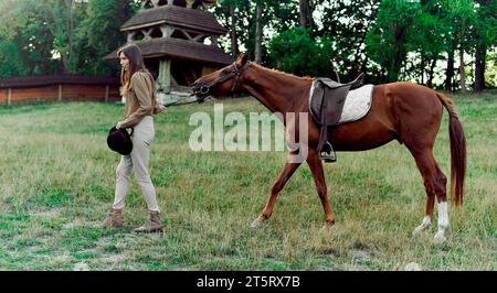 Junge Jockey-Mädchen, die das braune Sattelpferd an den Zügeln auf einem Rasenfeld anführt. Pferdeschule, Reitzentrum, Reitunterricht. Pferdesport nimmt ho Stockfoto