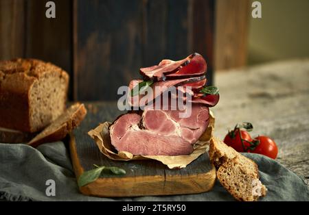 Nahaufnahme eines appetitlichen Schinkenstücks mit frischem Basilikum, Tomaten und Brot, serviert auf einem Holzbrett. Ästhetische Lebensmittelpräsentation. Konzept der Mutter Stockfoto