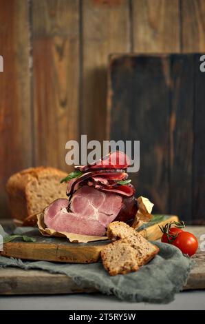 Ein Stück köstlicher Schinken, kombiniert mit Basilikumblättern, Reifen Tomaten und frischem Brot, elegant auf einer rustikalen Holzplatte angeordnet. Abbildung für die Lebensmittel Stockfoto