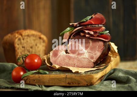 Ein großes Stück saftigen Schinken mit zwei Reifen Tomaten und Basilikoblättern, die auf einem Holzteller angeordnet sind. Ein Stück Vollkornbrot im Hintergrund. Stockfoto