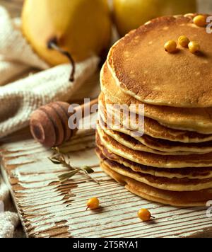 Nahaufnahme eines Haufens flauschiger Buttermilchpfannkuchen auf einer hellen Holzoberfläche, mit einem Paar Birnen in der Ferne, Sanddornbeeren und einem Stockfoto