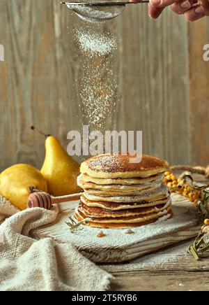 Puderzucker wird auf einen Haufen Buttermilchpfannkuchen mit einem Paar Birnen in der Ferne, einem kleinen Sanddorn-Zweig und einem Löffel für dri gestreut Stockfoto