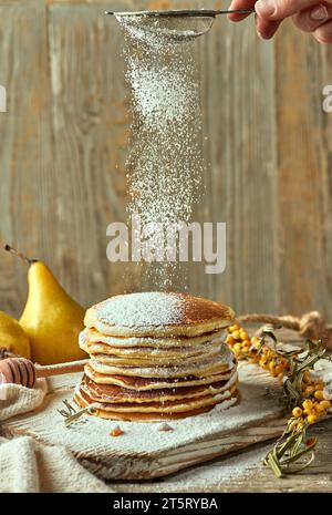 Vanillepfannkuchen auf einem hellen Holztisch, umgeben von einem Paar Birnen in der Ferne, einem kleinen Sanddornzweig und einem Löffel zum Gießen Stockfoto