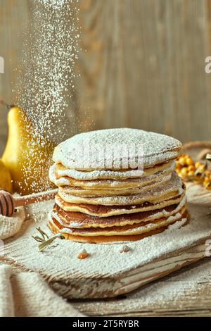 Nahaufnahme von hausgemachten Haferflockenpfannkuchen, mit Puderzucker bestäubt, auf einer hellen Holzplatte. Birnen und Sanddornbeeren im Hintergrund Stockfoto