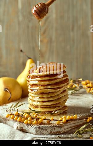 Honig fließt von einem Holzlöffel auf einem wunderschön präsentierten Stapel hausgemachter Pfannkuchen auf einem hellen Holzbrett mit einem Zweig Sanddorn und Stockfoto