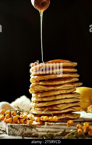 Bild von hausgemachten Pfannkuchen, die auf einem hellen Holzbrett mit einem Sanddornzweig angeordnet sind. Honig fließt aus einem Holzlöffel. Kontrastreiche Farbgebung Stockfoto