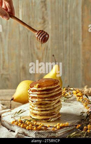 Klassische amerikanische Pfannkuchen auf einer hellen Holzplatte mit Sanddornzweigen und gelben Birnen. Honig fließt aus einem Holzlöffel. R Stockfoto
