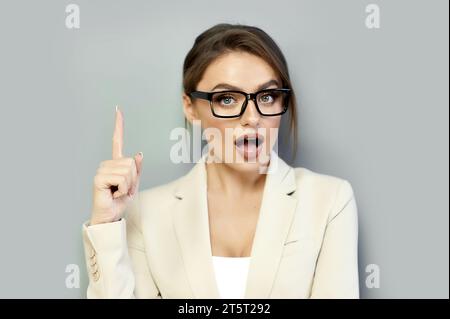 Aufgeregt attraktive dunkelblonde Frau in schwarz gerahmter Brille und beigefarbener Jacke, die auf freien Kopierraum auf grauem Hintergrund zeigt. Modell für Produktanzeige Stockfoto