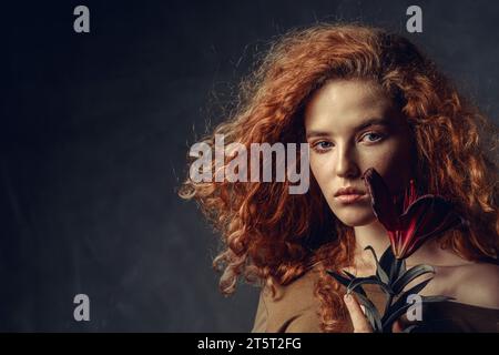 Kunstporträt einer schönen jungen rothaarigen Frau mit durchdringendem Blick und rotem Haar in Bewegung, die eine dunkelburgunderrote Lilienblume hält. Kopierbereich. Stockfoto