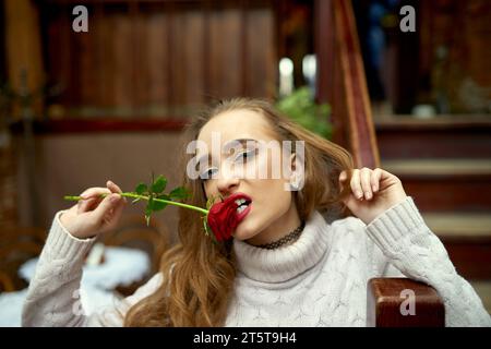 Ein junges Mädchen in einem weißen Pullover beißt eine Geschenkrose, um ihren verliebten Freund am Valentinstag zu verführen. Beißende rote Rose in das Blütenblatt. Lustige romantische Mutter Stockfoto