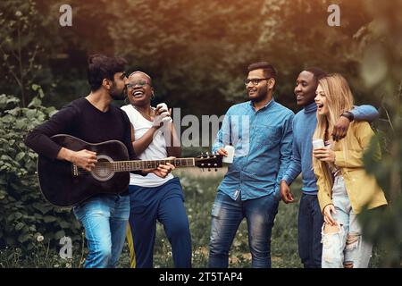 Ein junger Mann spielt Gitarre in einer multiethnischen Gruppe von fünf Personen. Freunde, die bei Sonnenuntergang in einem Park Spaß haben. Spontane Stau-Sitzung. Abhängen, Stockfoto