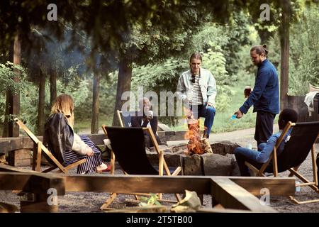 Multiethnische Gruppe von Freunden, die am Lagerfeuer rumhängen und Bier trinken. Ein Mann gießt Feuerzeugflüssigkeit in ein Feuer. Bei Sonnenuntergang abhängen. Aktive Lebensläufe Stockfoto