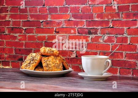 Ein Teller mit einem Haufen Shortbread-Kekse mit Samen und Erdnüssen auf einem Holztisch, daneben eine Tasse heißen Tee auf einer Untertasse. Nahaufnahme. Stockfoto