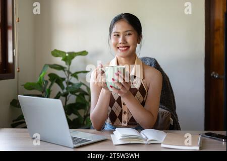 Eine charmante und glückliche asiatische Frau in lässiger Kleidung lächelt in die Kamera, während sie ihren Morgenkaffee an ihrem Schreibtisch in ihrem Heimbüro genießt. Stockfoto