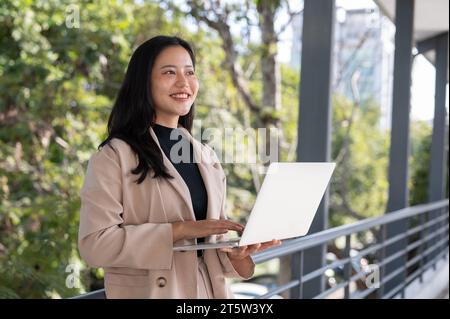 Eine attraktive asiatische Geschäftsfrau, die auf einem Skywalk in der Stadt an ihrem Laptop arbeitet. Städtisches Leben, Menschen und Technologiekonzepte Stockfoto