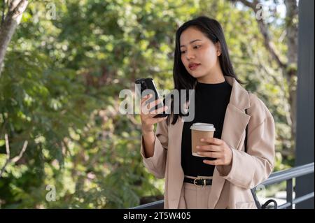 Eine wunderschöne asiatische Geschäftsfrau, die Millennials ist, reagiert auf Nachrichten auf ihrem Handy, während sie auf einem Skywalk läuft. Städtisches Leben, Menschen und Technologie Co Stockfoto