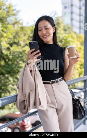 Eine wunderschöne asiatische Geschäftsfrau der Millennials reagiert auf Nachrichten auf ihrem Handy auf einem Skywalk. Städtisches Leben, Menschen und Technologiekonzepte Stockfoto