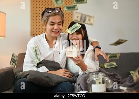 Glückliches, junges Paar, das Hausmodell hält, auf der Couch sitzt, mit Dollarscheine, die von oben fallen Stockfoto