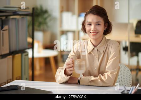 Lächelnde junge Geschäftsfrau, die eine Tasse Tee trinkt, wenn sie am Schreibtisch arbeitet Stockfoto