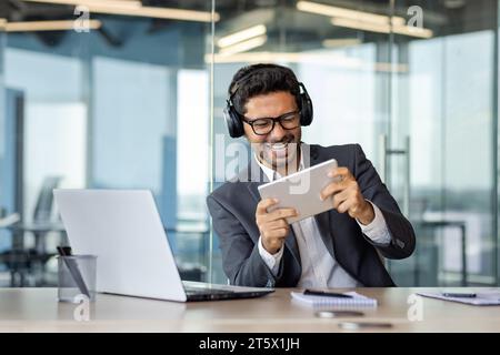 Ein Mann in Business-Anzug und Kopfhörern spielt ein Spiel auf einem Tablet-Computer im Büro, ein araber lächelt zufrieden mit seiner Ruhe, der Chef arbeitet mit einem Laptop. Stockfoto