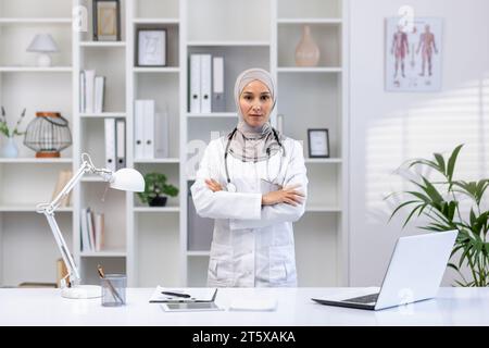 Porträt der ernsthaften selbstbewussten erfolgreichen weiblichen Ärztin im Hijab im weißen medizinischen Mantel, die Kamera mit gekreuzten Armen betrachtet, Klinikarbeiter, die im Büro medizinischen Raum arbeiten. Stockfoto