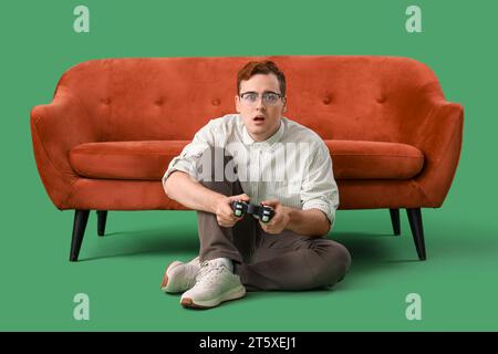 Schockierter junger Mann, der Videospiel in der Nähe des roten Sofas auf grünem Hintergrund spielt Stockfoto