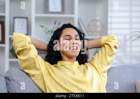Nahaufnahme einer erfolgreichen lächelnden und zufriedenen Frau mit Händen hinter dem Kopf mit geschlossenen Augen lächelnd und glücklich sitzend auf dem Sofa im Wohnzimmer zu Hause im Haus. Stockfoto