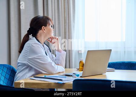 Selbstbewusste ernste reife Ärztin, die mit Laptop am Tisch sitzt Stockfoto