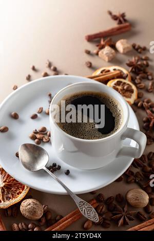 Schwarzer Kaffee in weißer Tasse. Auf beigefarbenem Hintergrund sind Kaffeebohnen, Zimtstangen, Anis, Nelken, Muskatnuss, Pfeffer, brauner Zucker und getrocknetes oder Stockfoto