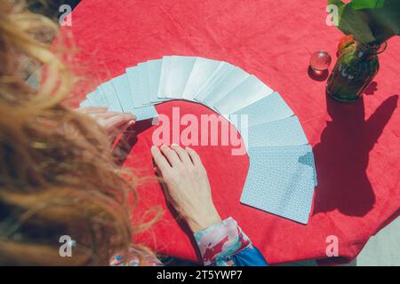 Ansicht von oben und hinten von nicht erkennbaren kaukasischen Frauenhänden, die Tarotkarten von Deck auf Tisch mit roter Tischdecke und Kopierraum nehmen Stockfoto