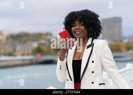 Elegante afroamerikanische Geschäftsfrau, die draußen neben einem städtischen Fluss eine Sprachnachricht schickt Stockfoto