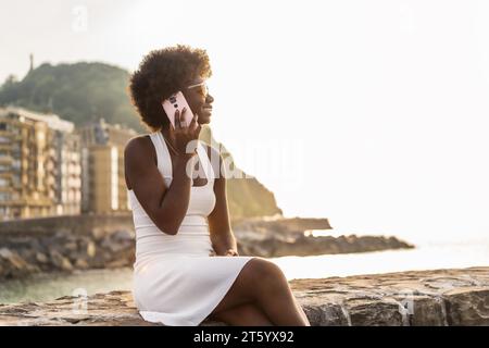 Junge Afro-Frau, die auf einer Promenade sitzt und mit dem Handy spricht und die Aussicht auf den Sonnenuntergang genießt Stockfoto