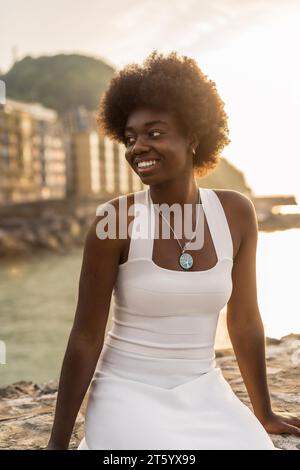 Vertikales Porträt einer glücklichen Schönheit afrikanerin in weißem Sommerkleid, die bei Sonnenuntergang draußen am Meer sitzt Stockfoto