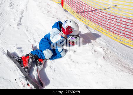 Der Vorschuljunge in Helm, Brille, Ski und Winteranzug lernt Ski, fällt und liegt auf dem Schnee. Das müde Kind legte sich auf den Schnee, um sich auszuruhen. Stockfoto