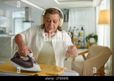 Ältere Frau hört Podcasts in Kopfhörern, wenn sie zu Hause Kleidung bügelt Stockfoto