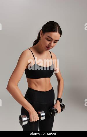 Motivierte und starke Sportlerin in schwarzer aktiver Kleidung, die mit Kurzhanteln vor grauem Hintergrund trainiert Stockfoto