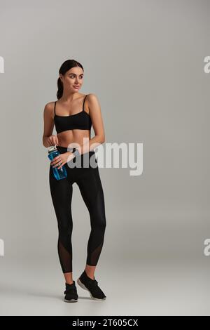 Sportlerin in Leggings, Crop-Top und Sneakers, die Flasche mit Wasser auf grauem Hintergrund hält Stockfoto