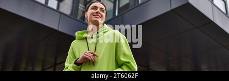 Niedriger Blickwinkel auf eine glückliche Sportlerin in einem übergroßen Hoodie, die draußen lächelnd ist, horizontales Banner Stockfoto