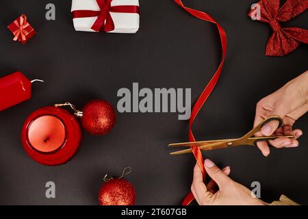 Zugeschnittene Ansicht einer Frau, die ein Band mit einer Schere in der Nähe von Kugeln schneidet, und Weihnachtsdekor auf Schwarz Stockfoto