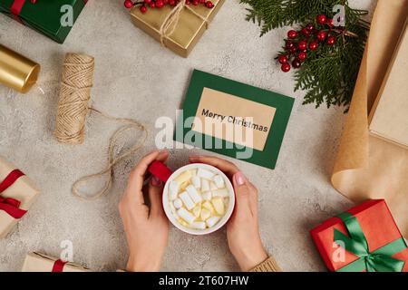 Zugeschnittene Ansicht einer Frau, die heiße Schokolade mit Marshmallows in der Nähe der Grußkarte „Merry Christmas“ hält Stockfoto